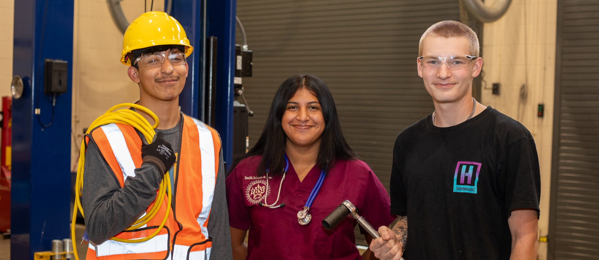 Three high school students stand together, representing different career paths. On the left, a student wears a yellow hard hat, safety glasses, and an orange safety vest with a coiled yellow cable over their shoulder, representing construction or engineering. In the center, a female student in burgundy scrubs wears a stethoscope, representing healthcare. On the right, a student in a black t-shirt with a logo holds a long tool, possibly representing a trade or technical field. They are in an industrial-looking setting with equipment visible in the background. The image showcases vocational education options available to high school students.