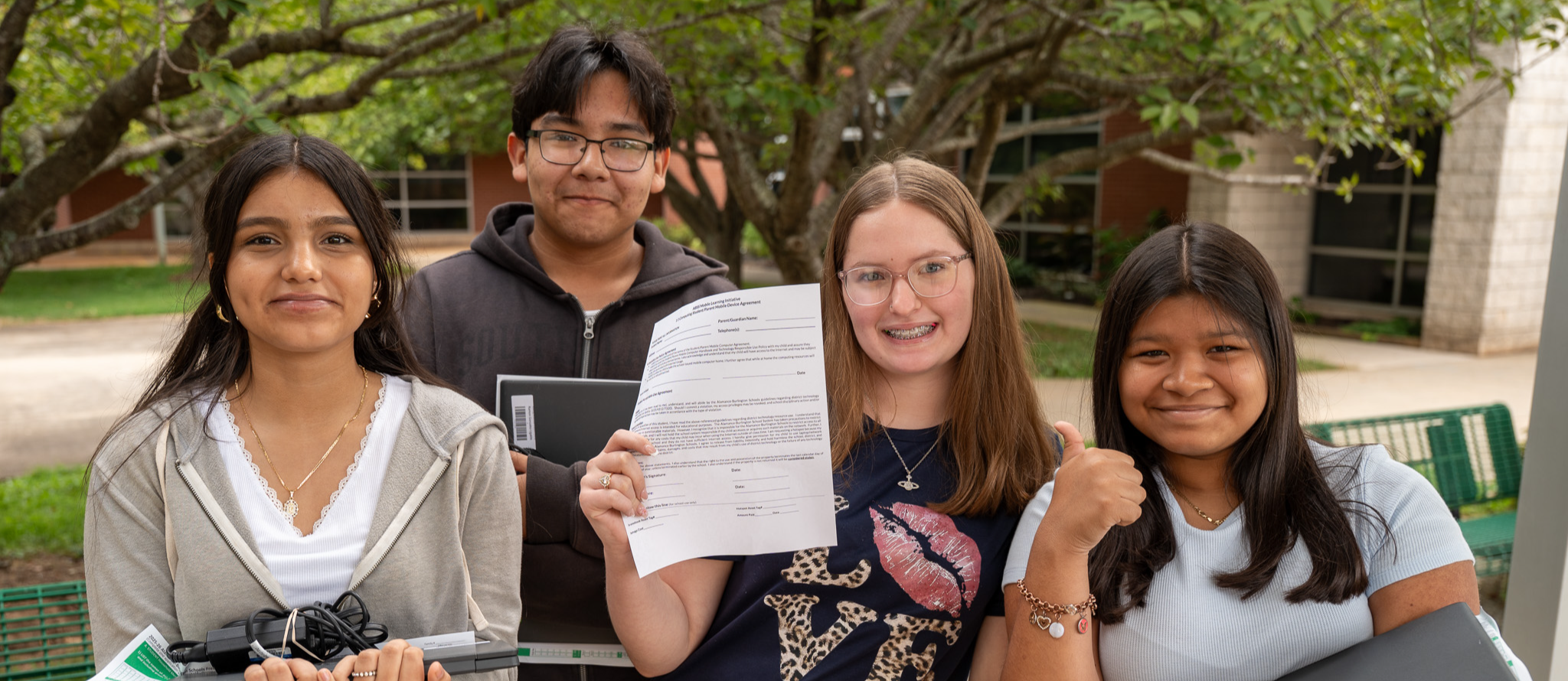 Four high school students holding Chromebooks and computer accessories, one holding up a student contract, on campus with trees behind them
