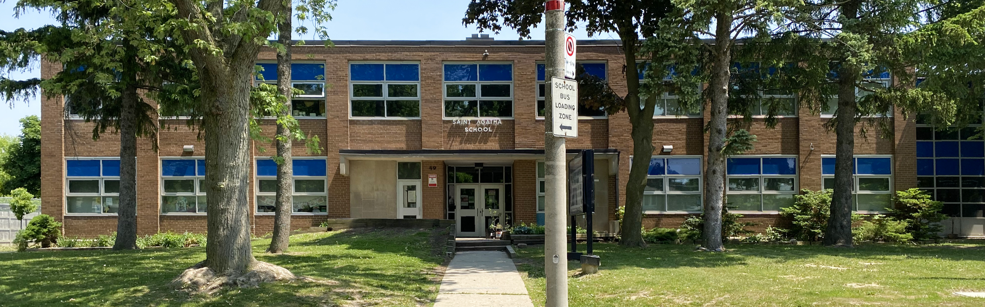 Photo of the St. Agatha Catholic School building, showing a tan brick building with many windows, which feature both clear and blue glass panels, the school name is displayed in white above the main entrance, and is surrounded by many trees, grass, and a school signboard. 