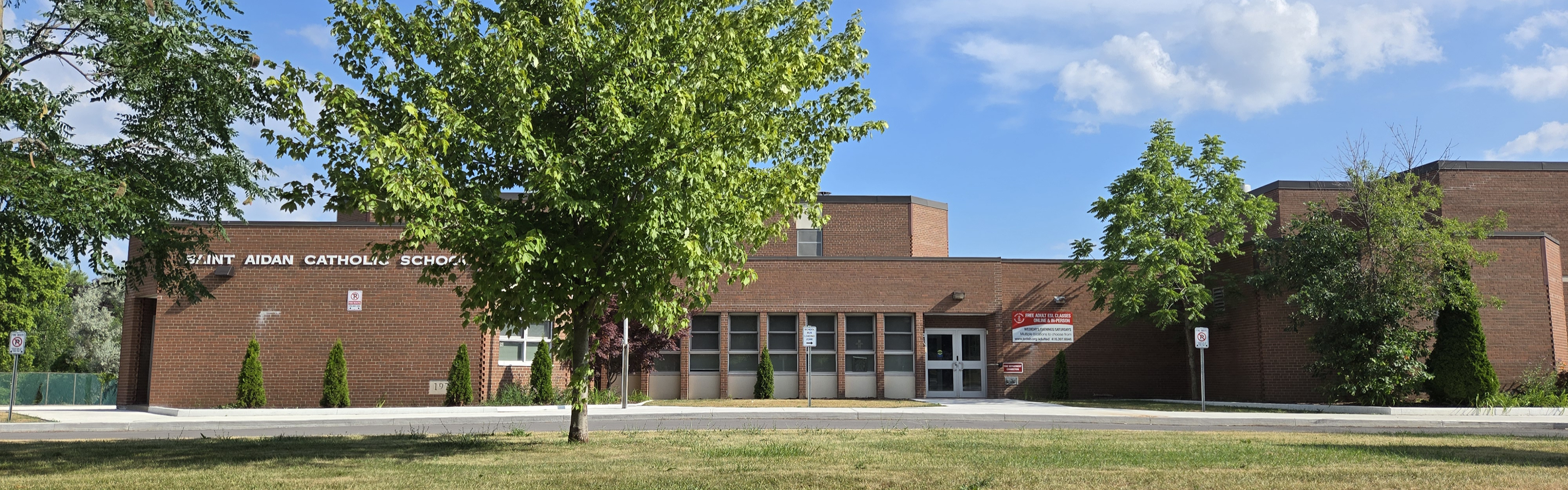 A photo of the St. Aidan Catholic School building, showing a brown brick building with the school name displayed in white, trees and grass surrounding it, looking onto the driveway. 