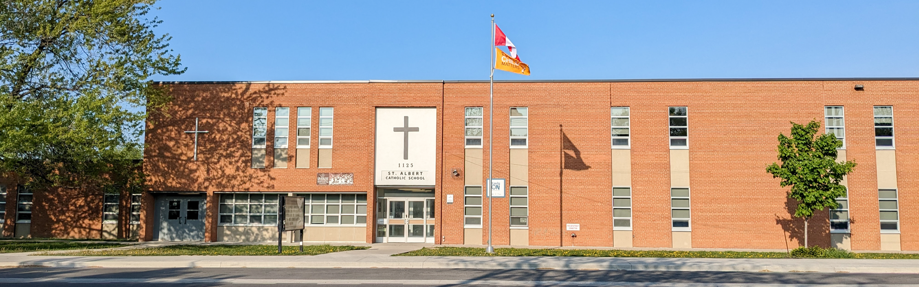 A photo of the St. Albert Catholic School building, showing a red brick building with narrow windows, the school name and a large black cross sit above the entrance on a white portion of the wall. An additional thin white cross sits on the red brick portion to the left side of the wall within the shadows of a nearby tree. The school bulletin is planted in the grass as two raised flags sit atop the flagpole flapping in the wind. 