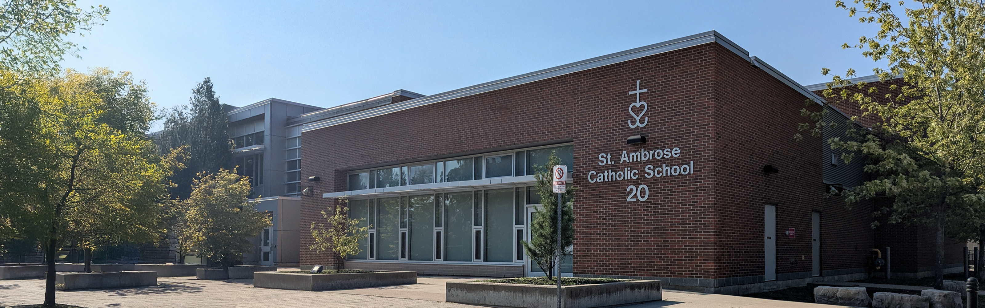 Photo of the St. Ambrose Catholic School building, showing a modern red brick building with large windows and the school name displayed in white on the wall, surrounded by trees and landscaped planters. 