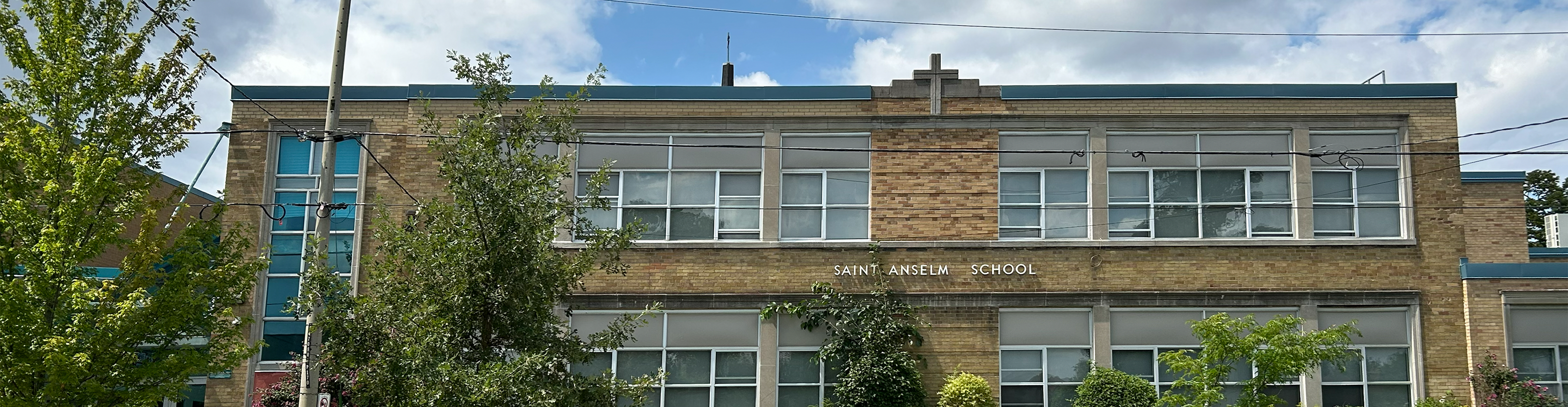The front of the St. Anselm Catholic School building, showing a yellow brick building looking out onto a residential road, bordered by trees and greenery