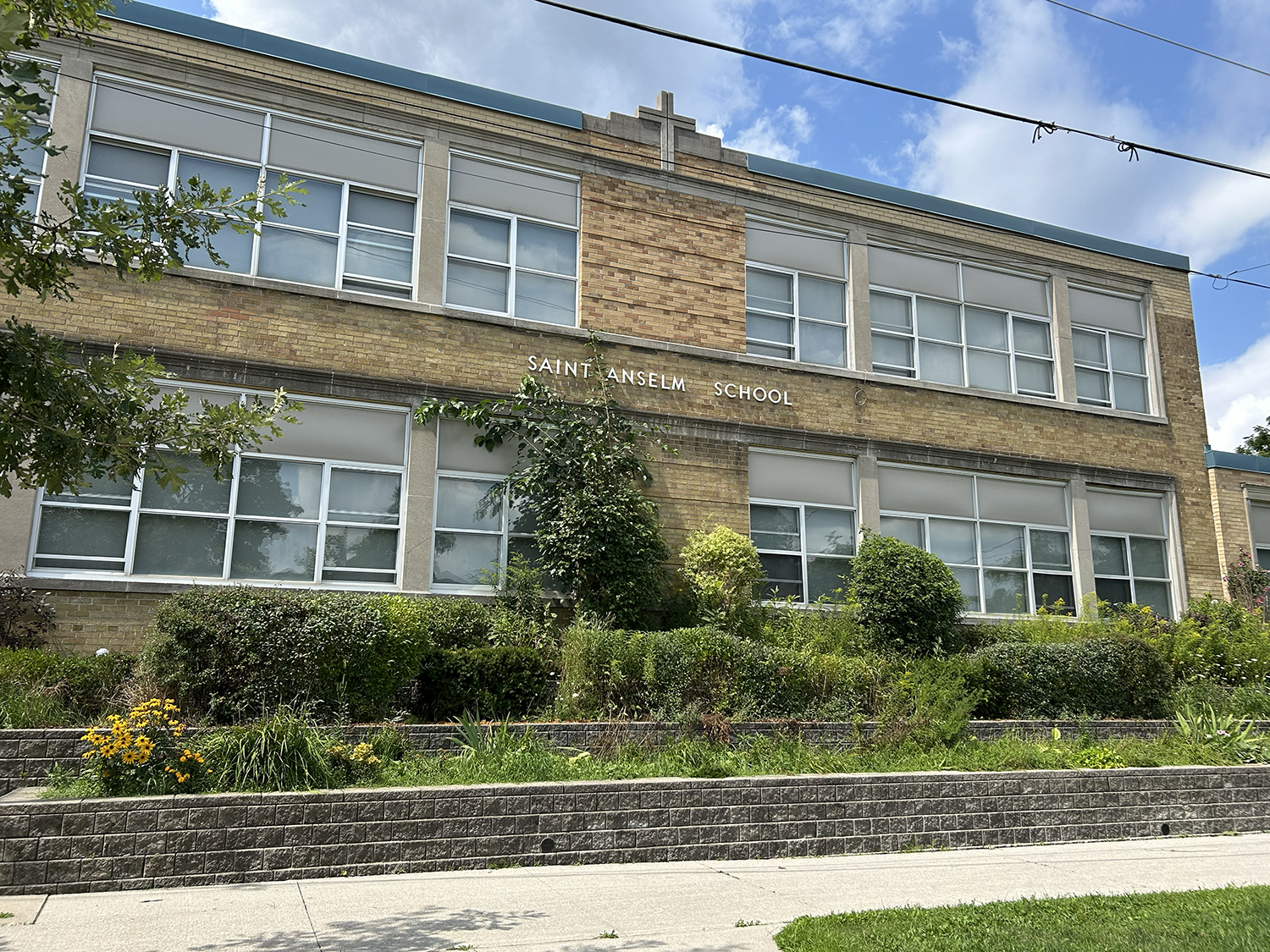 The front of the St. Anselm Catholic School building, showing a yellow brick building looking out onto a residential road, bordered by trees and greenery
