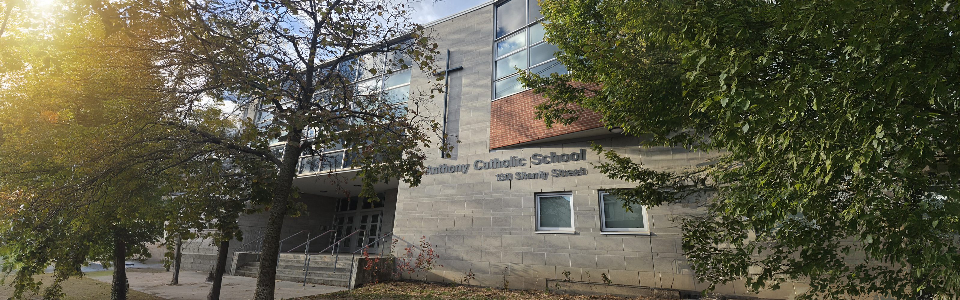 Photo of the St. Anthony Catholic School building, showing a modern gray stone building with a large cross above the entrance, the school name displayed in grey, large glass windows, steps leading up to the main doors and surrounding trees.   