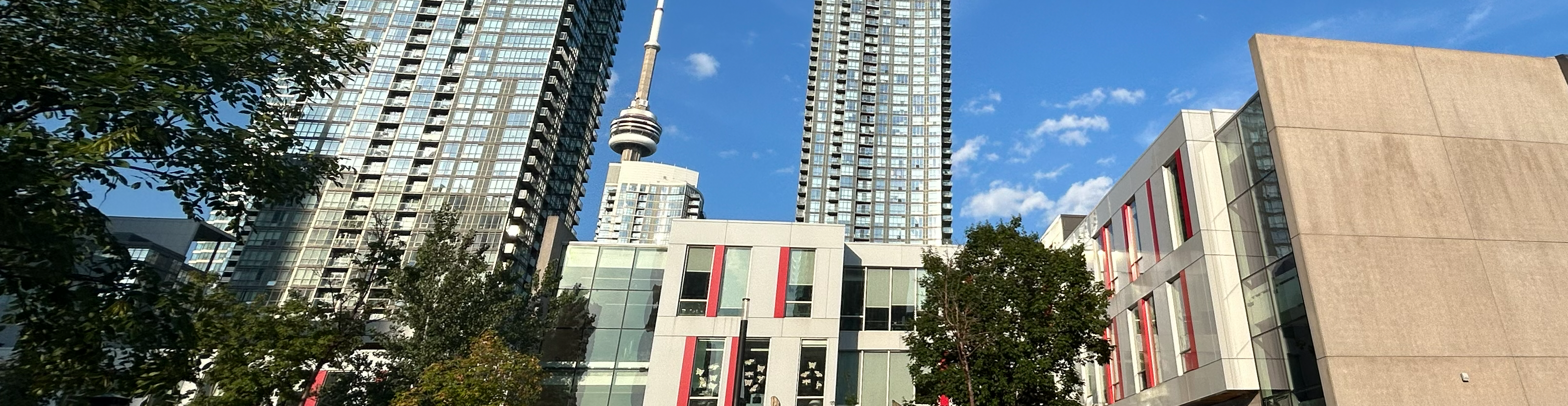 Photo of the Bishop Macdonell school building as part of the larger Canoe Landing Community Recreation Centre, nestled between buildings in downtown Toronto, with the CN Tower visible in the background and trees visible in the foreground