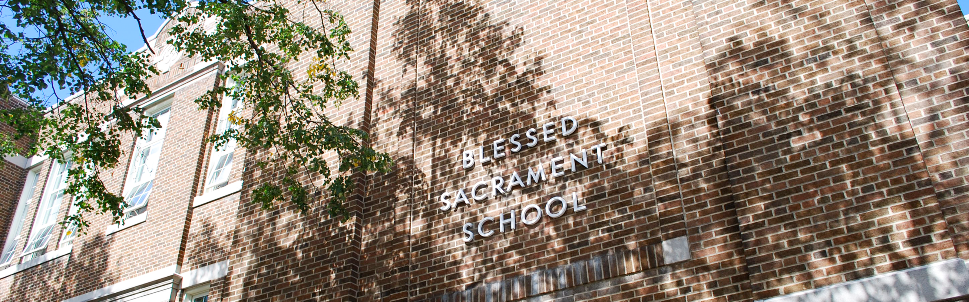 Photo of the Blessed Sacrament Catholic School building, showing a red brick building with large windows, the school name on the front in white, surrounded by shadows cast by nearby trees.
