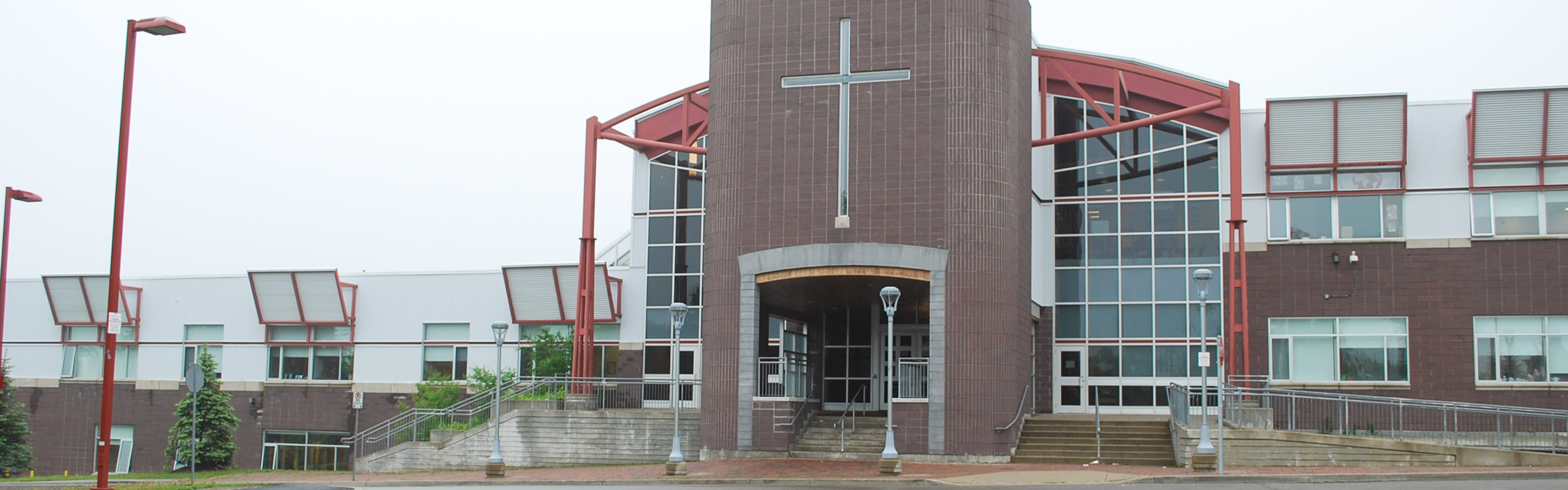 Photo of the Brebeuf College Catholic Secondary School building, showing a modern red brick building with large windows, red steel beams and a large cross above the main entrance.