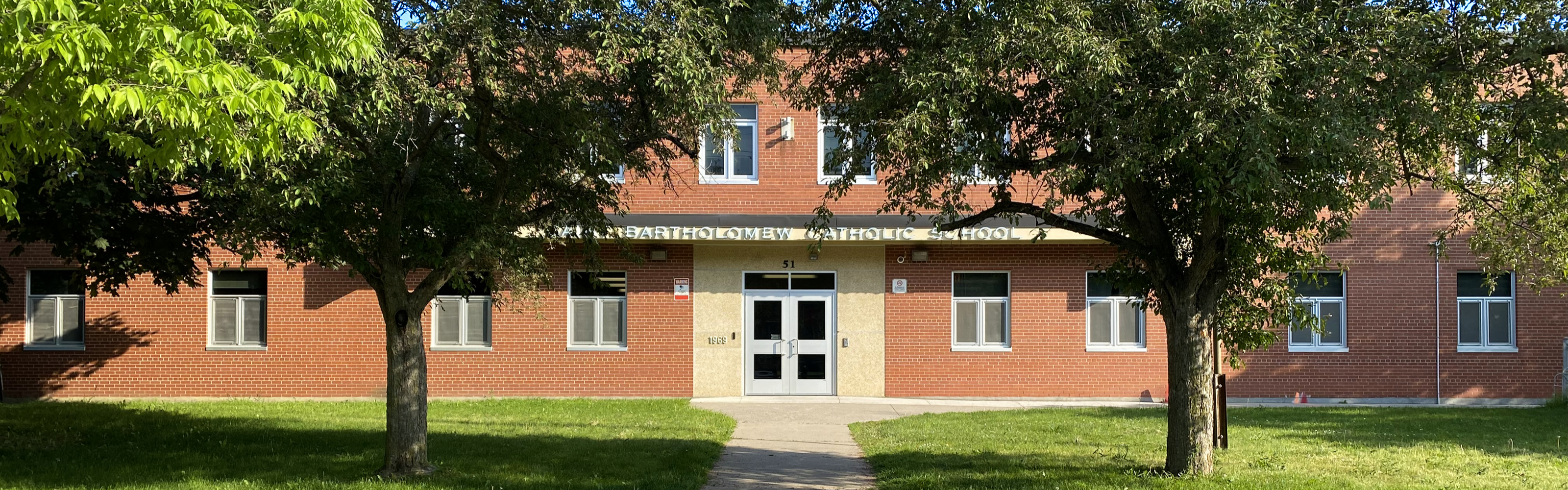 Photo of the St. Bartholomew Catholic School building, showing a red brick building with white framed windows, the school name displayed on a beige overhang above the entrance, framed by two large leafy trees on a green lawn.