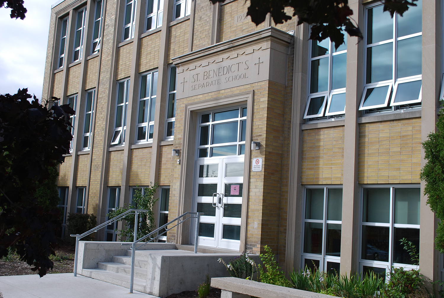 Photo of the St. Benedict School building, showing a beige school building surrounded by trees, looking out onto a busy main avenue
