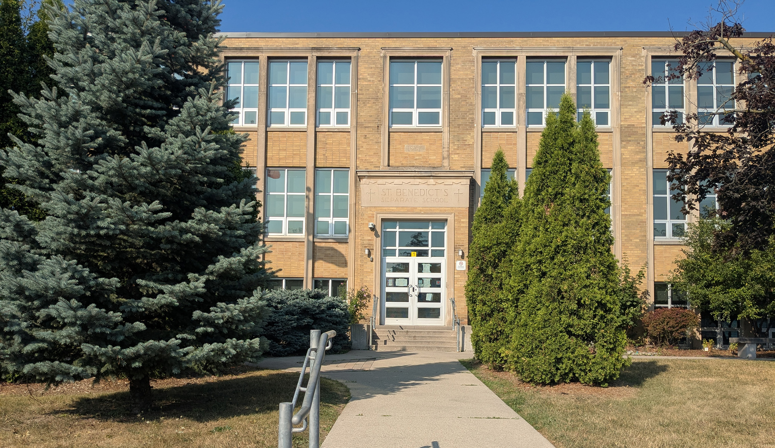 Photo of the front of the St. Benedict Catholic School building, showing a beige brick building bordered by trees and looking out onto a busy main road