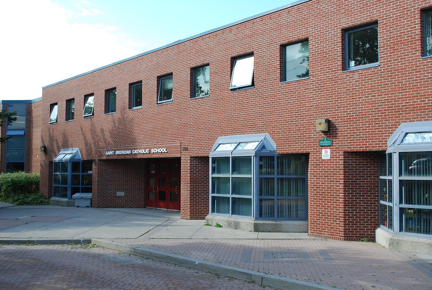 Photo of the St. Brendan Catholic School building, showing a red brick building bordered by tall trees, looking out onto a residential road