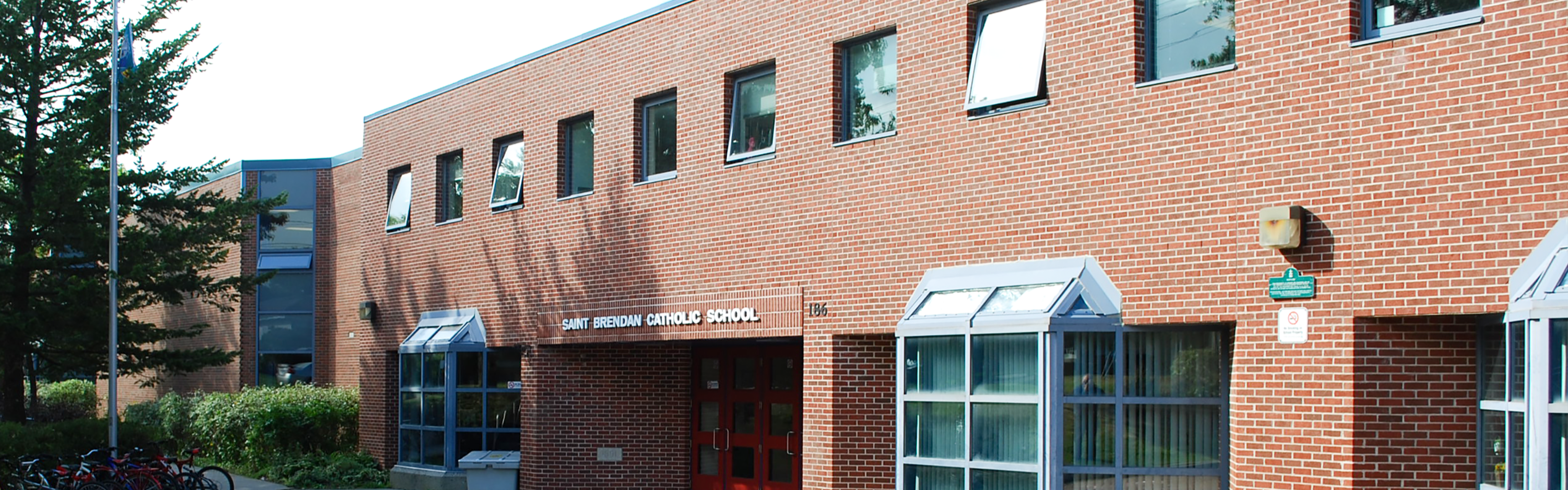 Photo of the St. Brendan Catholic School building, showing a red brick building with angled glass bay windows on the ground floor, red entrance doors, the school name displayed above the entrance in white, rows of upper windows, and green trees, bushes, a full bicycle rack, and a flagpole to the left of the entrance.