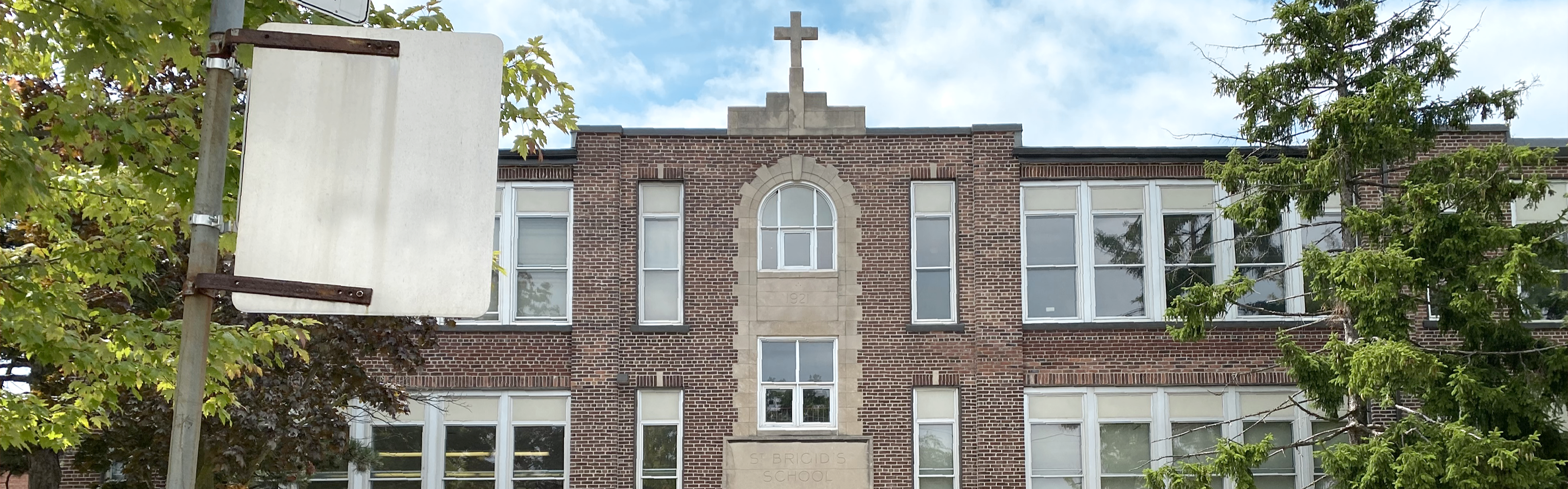 Photo of the St. Brigid Catholic School building, showing a traditional  red brick building with stone trim, an arched central window beneath a stone cross atop the roof, the school name carved above the entrance, with a large pine tree to the right.