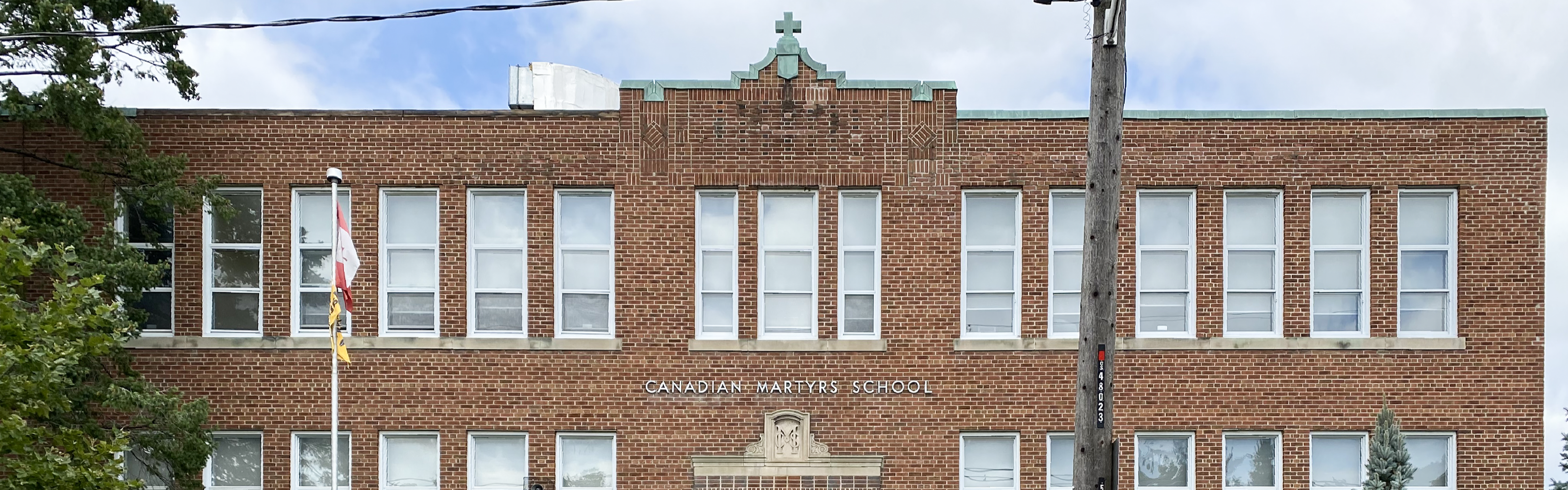 Photo of the Canadian Martyrs Catholic School building, showing a traditional red brick school building with white framed windows, a copper green trim featuring a small decorative cross in the center, and the school name displayed in white. 