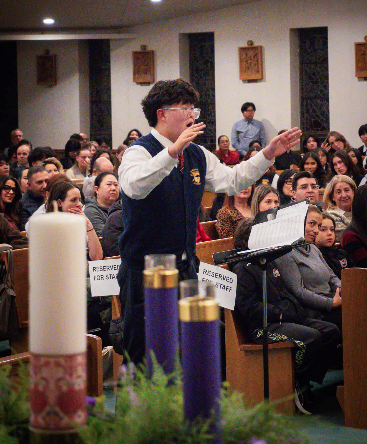 A young conductor gestures before a seated choir and audience in a church hall, with candles and sheet music in the foreground