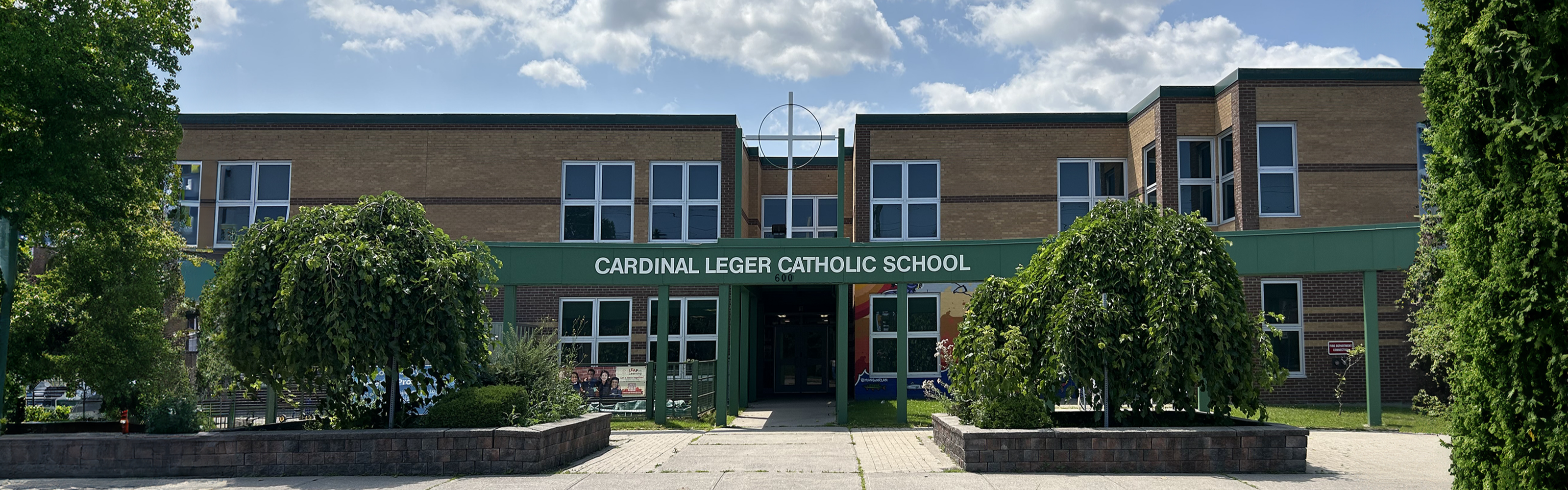 Photo of the Cardinal Léger Catholic School building, showing a modern brown and beige brick building with white windows, green steel beams, and a large white cross and the school's name above the entrance looking out onto the sidewalk, nestled between trees and bushes. 