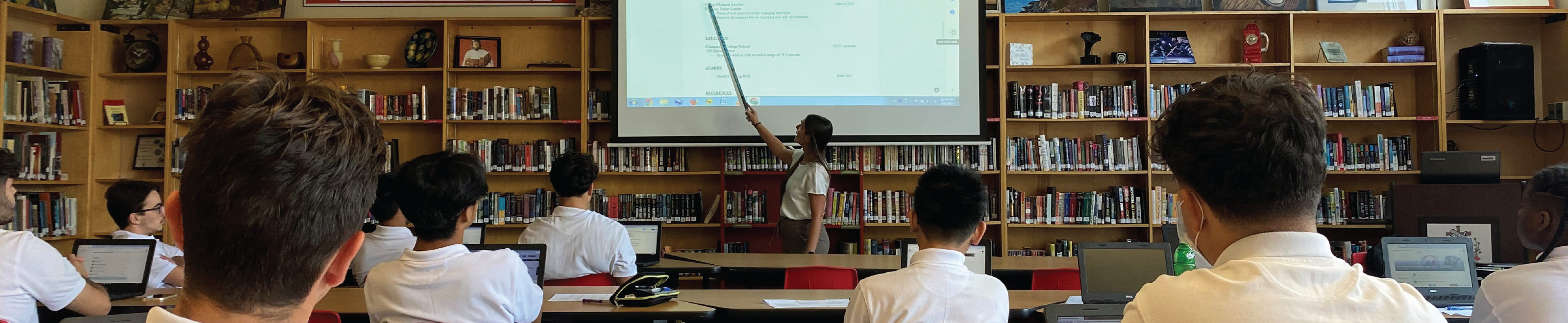 A female teacher pointing at the white screen while students are listening. 
