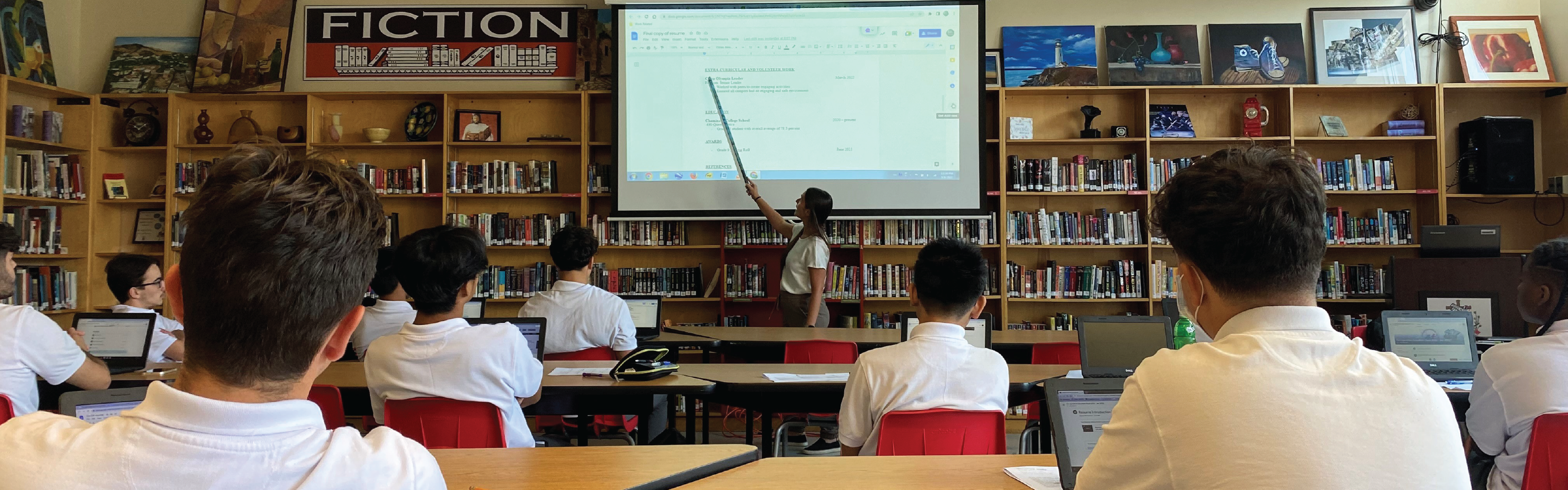 A female teacher pointing at the white screen while students are listening. 