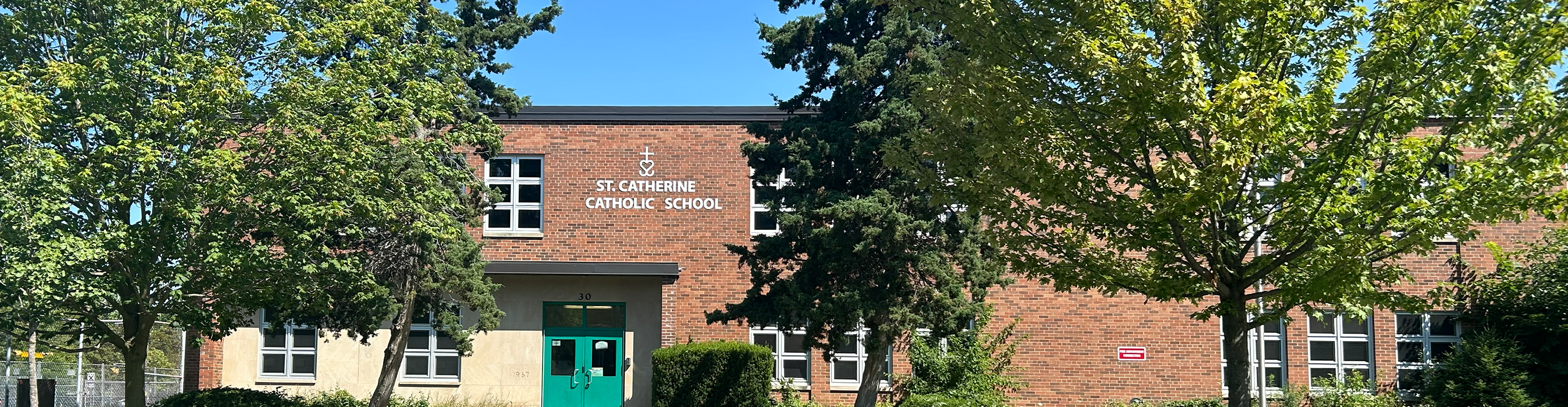 The front of the St. Catherine Catholic School building, showing the red brick building nestled between tall trees, with the school name in white above the front door