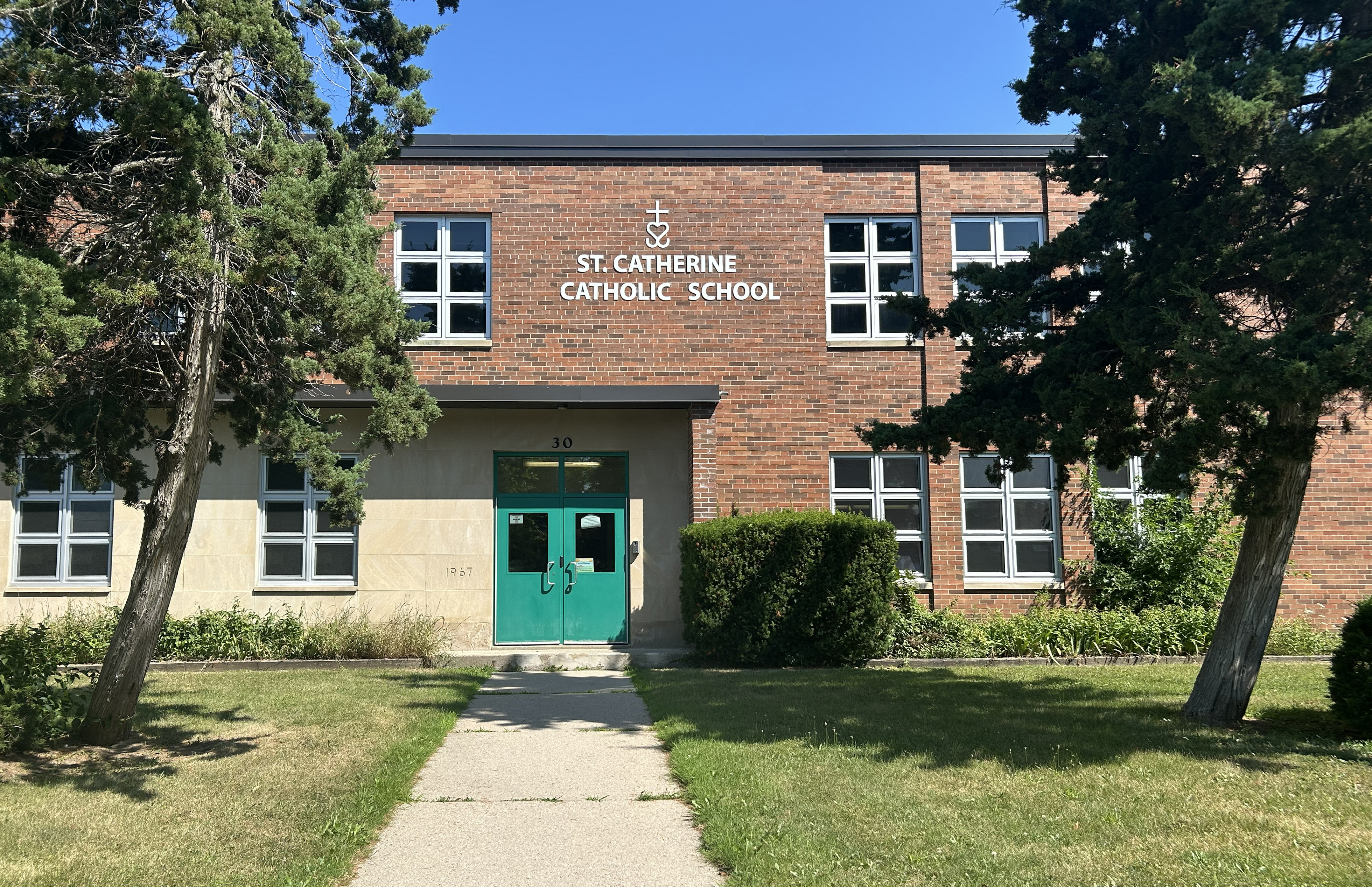The front of the St. Catherine Catholic School building, showing the red brick building nestled between tall trees, with the school name in white above the front door