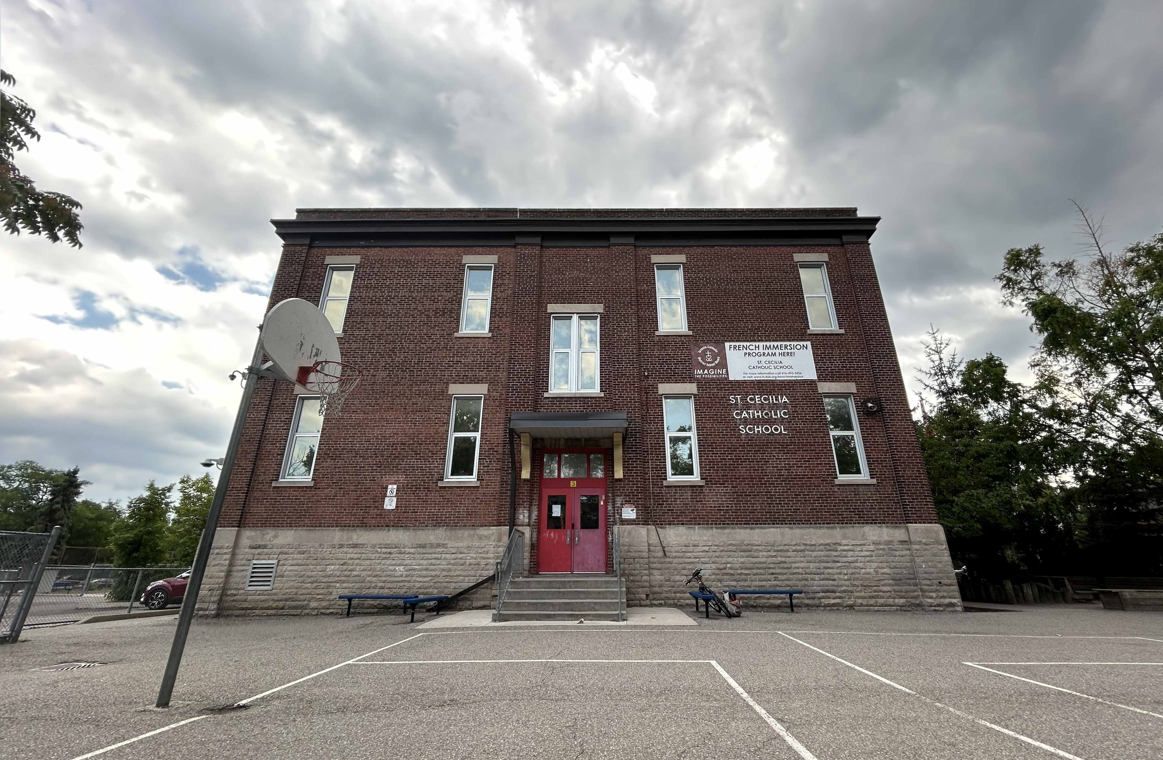 Photo of the St. Cecilia Catholic School building, showing a compact red brick and grey stone building looking out onto the school playground, with the school name in white on the front and a red painted door.