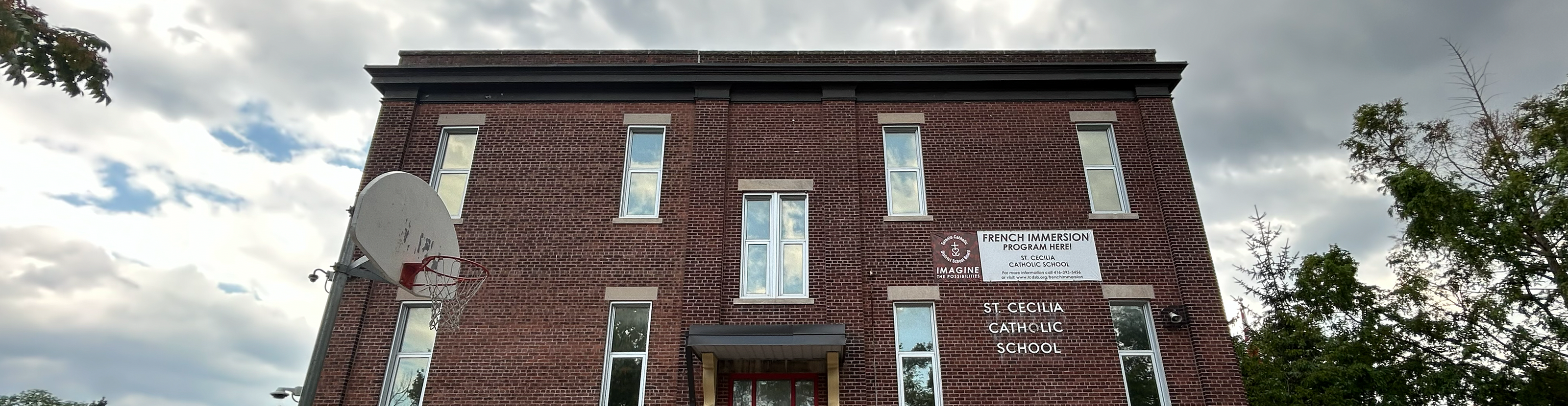 The front of the St. Cecilia Catholic School building, showing a compact dark red brick building with a red front door and the school name in white on the front