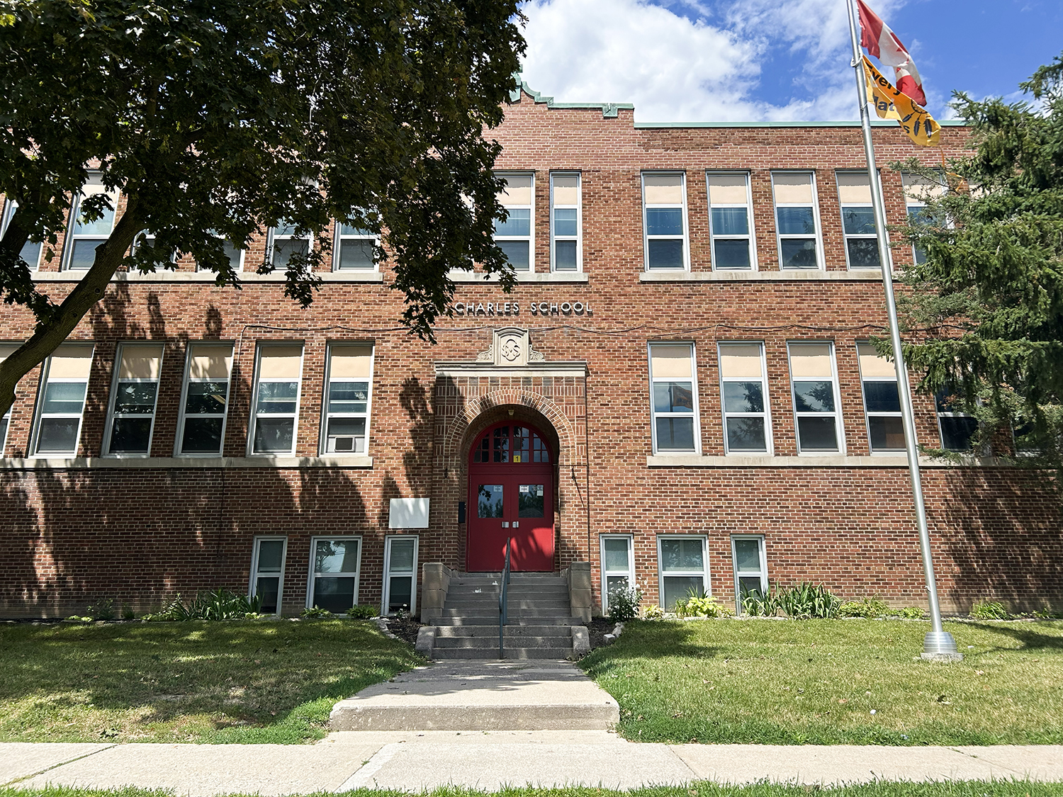 Photo of the St. Charles Catholic School building, showing a light red brick building with a red front door and the school's name in white above the door, surrounded by tall trees