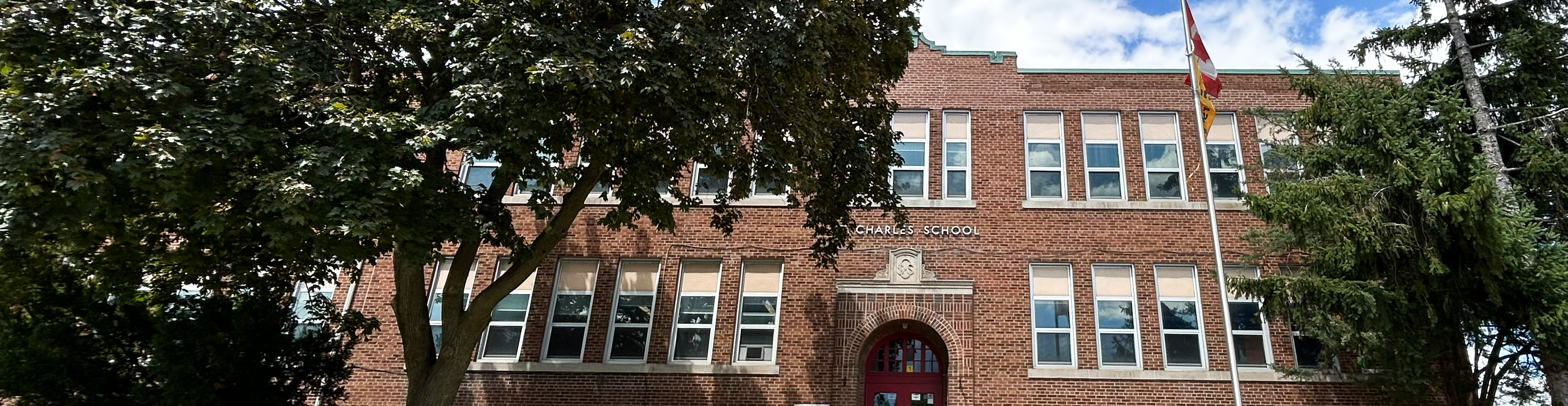 The front of the St. Charles Catholic School building, showing a red brick building bordered by tale trees, with a red front door with the school name in white above it