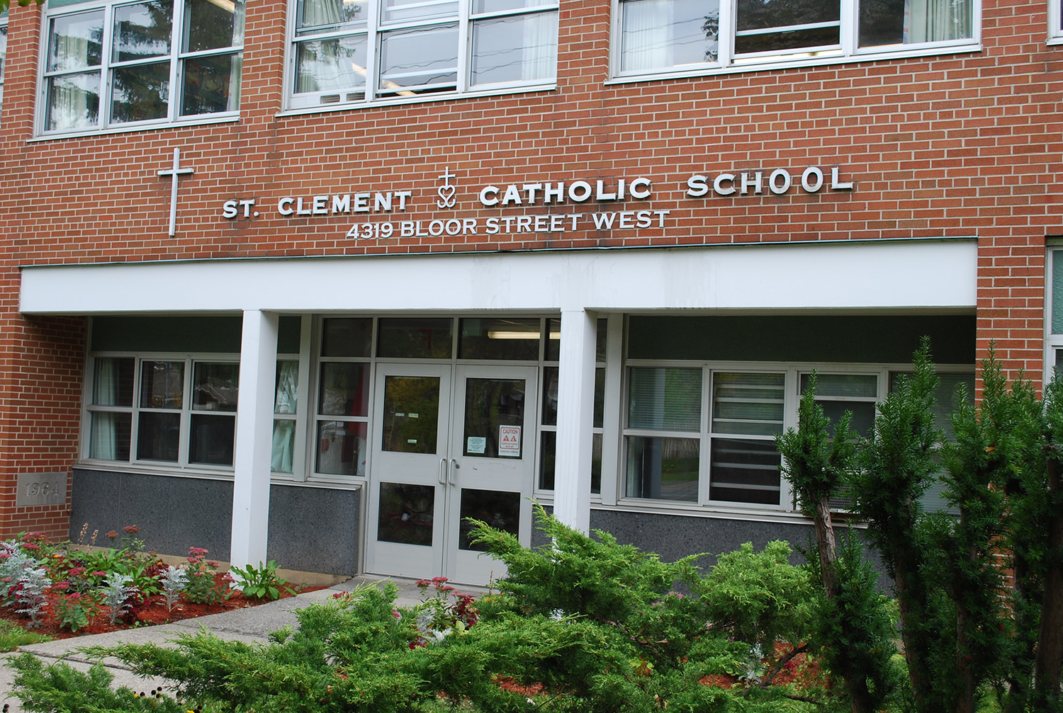 Photo of St. Clement Catholic School, a red brick building surrounded by some greenery looking out onto a busy main street