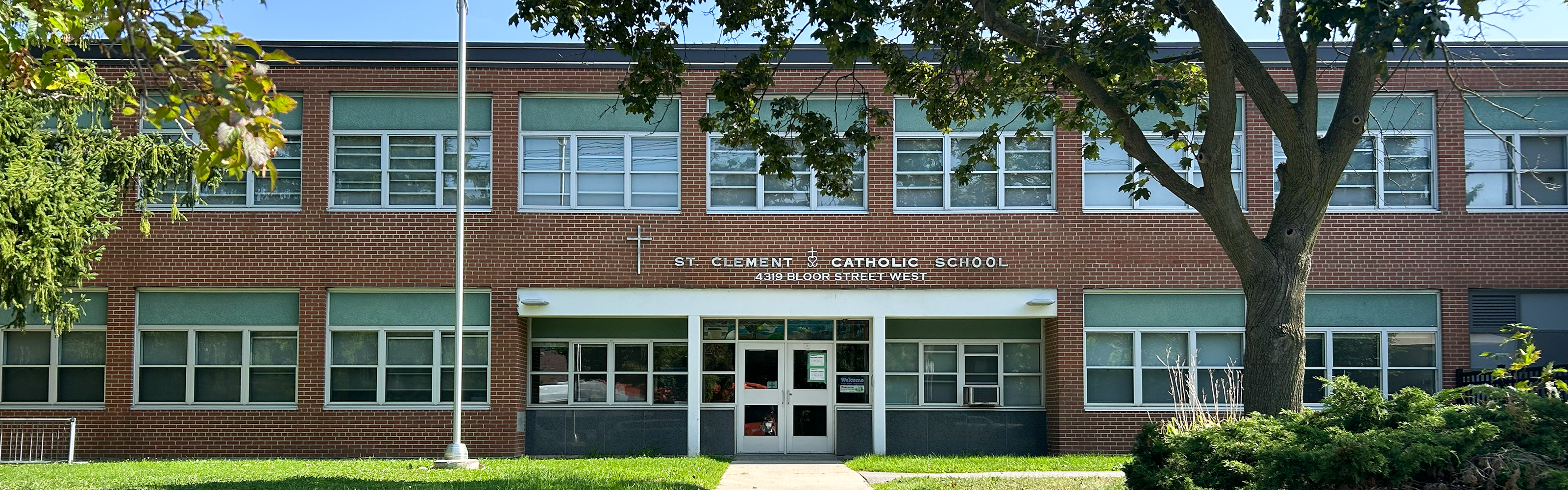 Photo of the St. Clement Catholic School Building, showing the red brick and glass building looking out onto the road, with the school name and address in white above the door, and bordered by trees