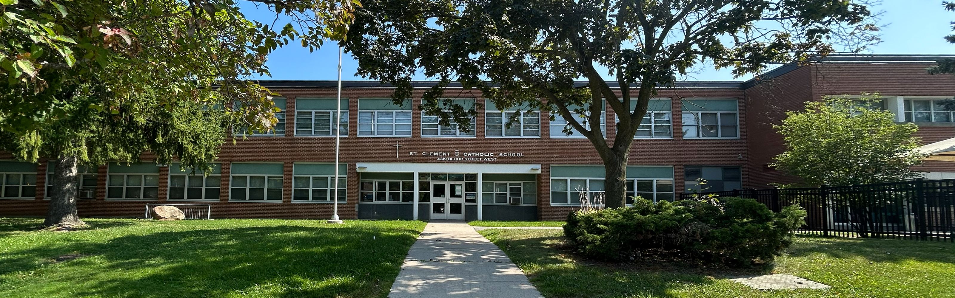 Photo of the St. Clement Catholic School Building, showing the red brick and glass building looking out onto the road, with the school name and address in white above the door, and bordered by trees