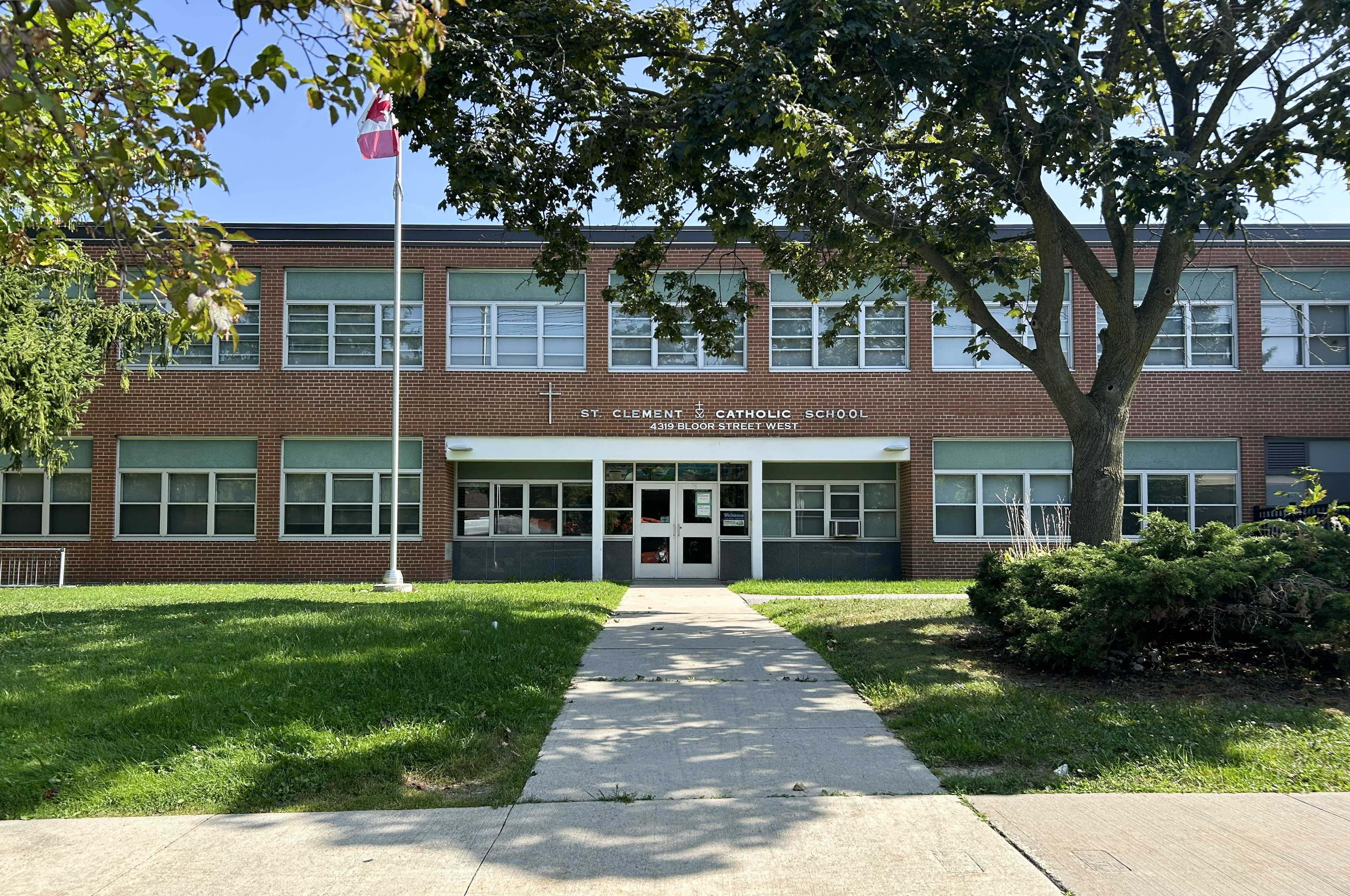 Photo of the St. Clement Catholic School Building, showing the red brick and glass building looking out onto the road, with the school name and address in white above the door, and bordered by trees