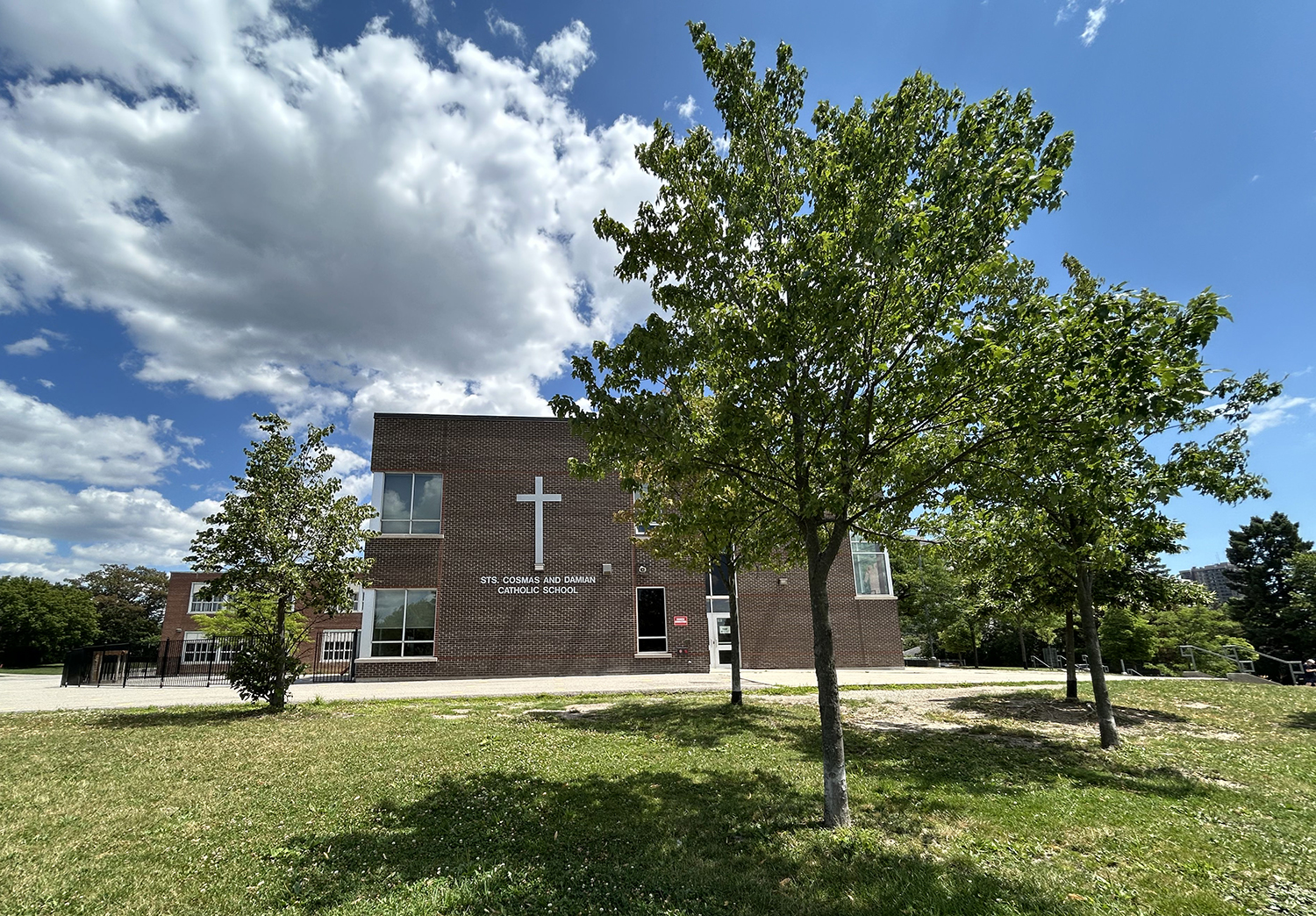 The front of the Sts. Cosmas and Damian Catholic School building, showing a compact red brick building situated in sprawling greenery and bordered by trees, with the school name in white on the front along with a large cross