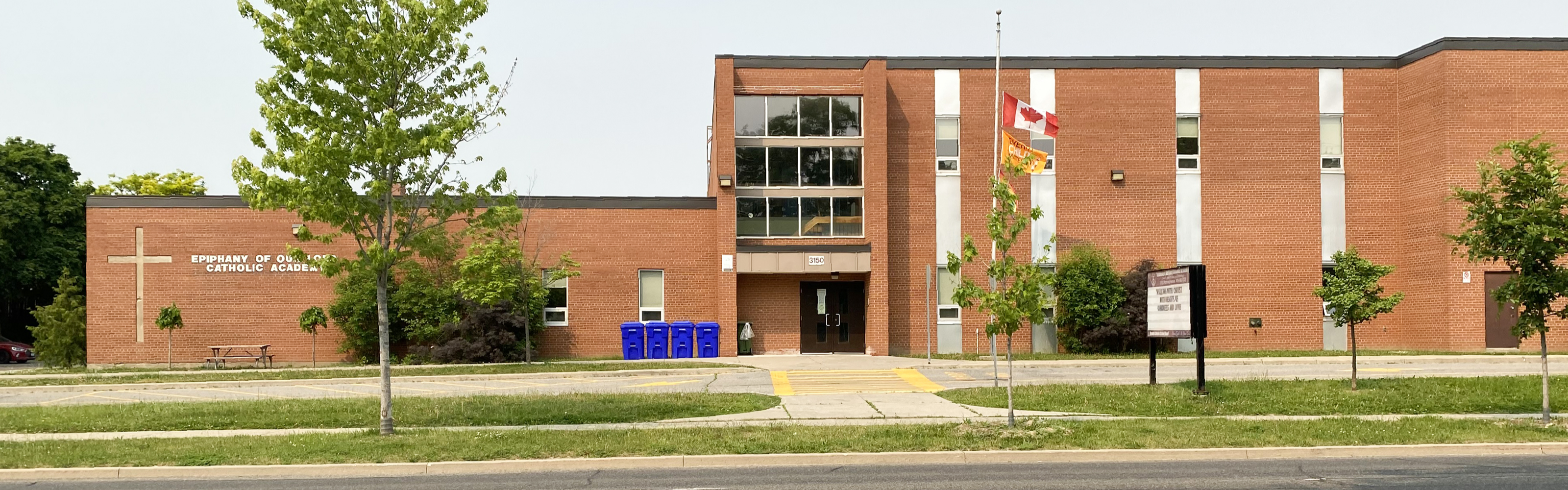 Photo of the Ephiphany of our Lord Catholic Academy building, showing a red brick building looking out onto the road, nestled between young trees, a school sign, with a large cross and the school name in white displayed on the left side of the building.