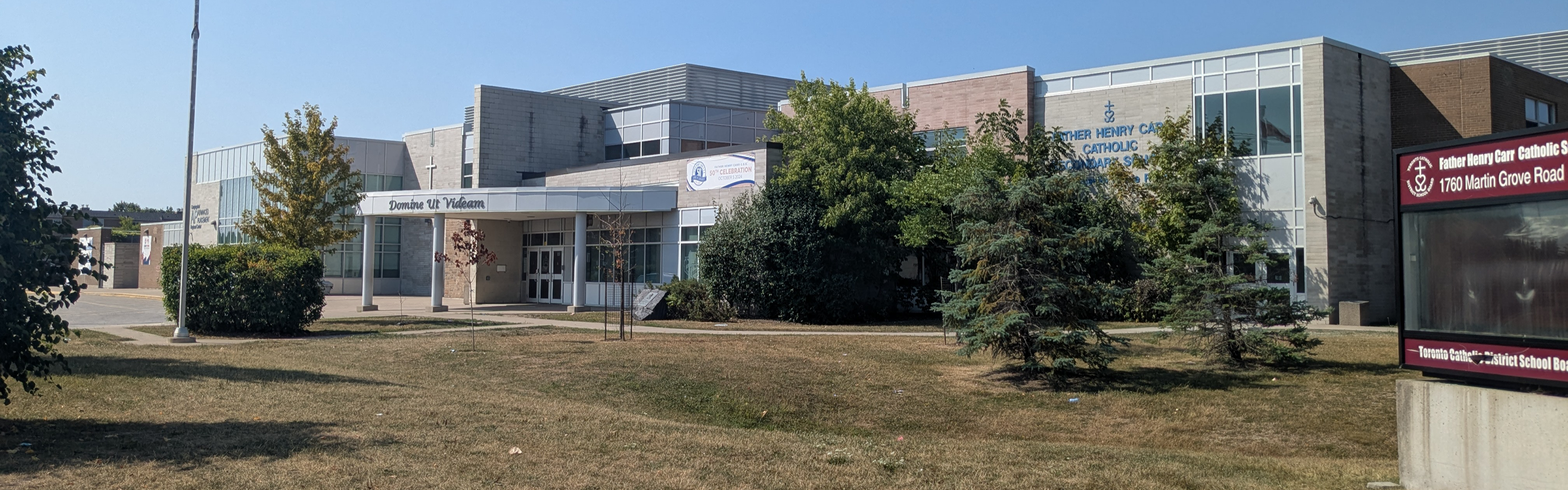 Photo of the Father Henry Carr Catholic Secondary School building, showing a pale brick building with large windows looking out onto the road and the school name in blue, nestled between a handful of trees, bushes, and grass with a digital school sign sitting on the grass to the left. 