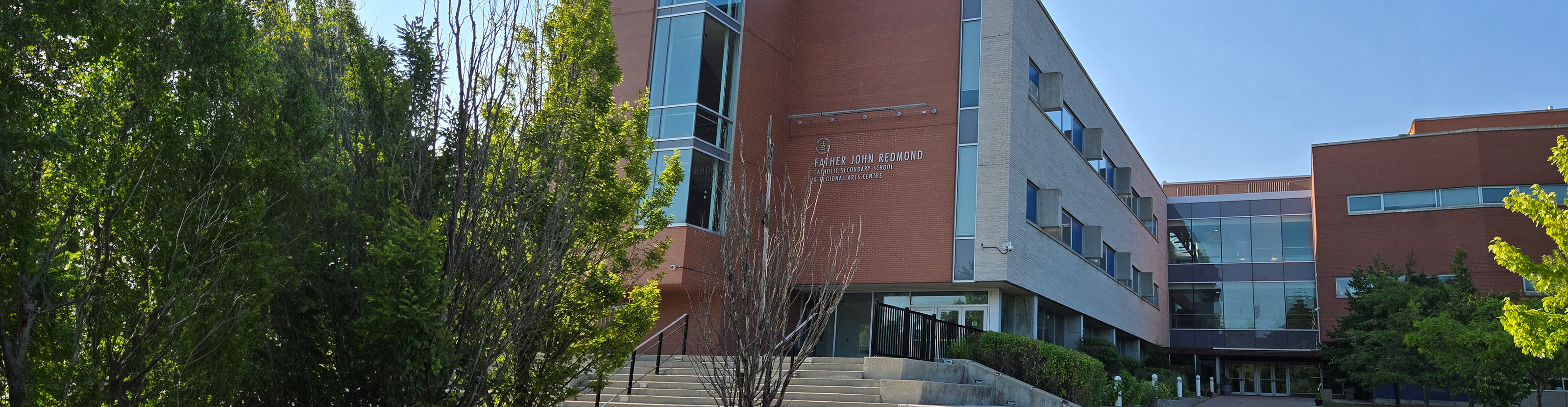Photo of the Father John Redmond Catholic Secondary School and Regional Arts Centre building, showing a modern red-and-gray building with large windows surrounded by many trees and bushes, the school name is displayed above the entrance, leading to a staircase.