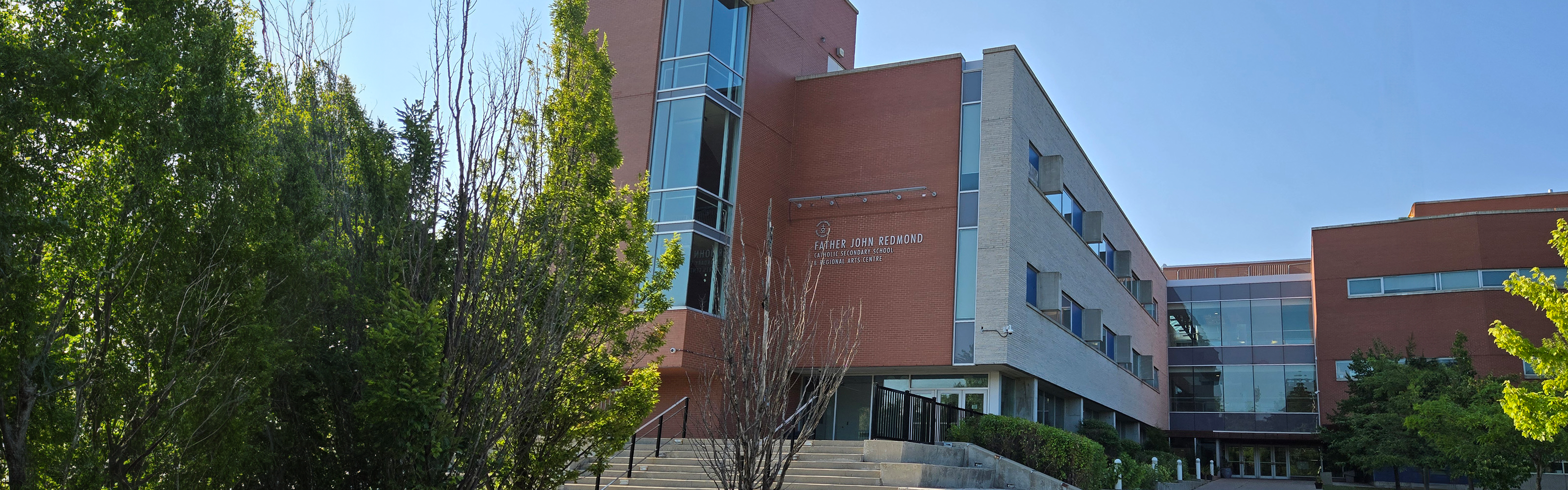 Photo of the Father John Redmond Catholic Secondary School and Regional Arts Centre building, showing a modern red-and-gray building with large windows surrounded by many trees and bushes, the school name is displayed above the entrance, leading to a staircase.