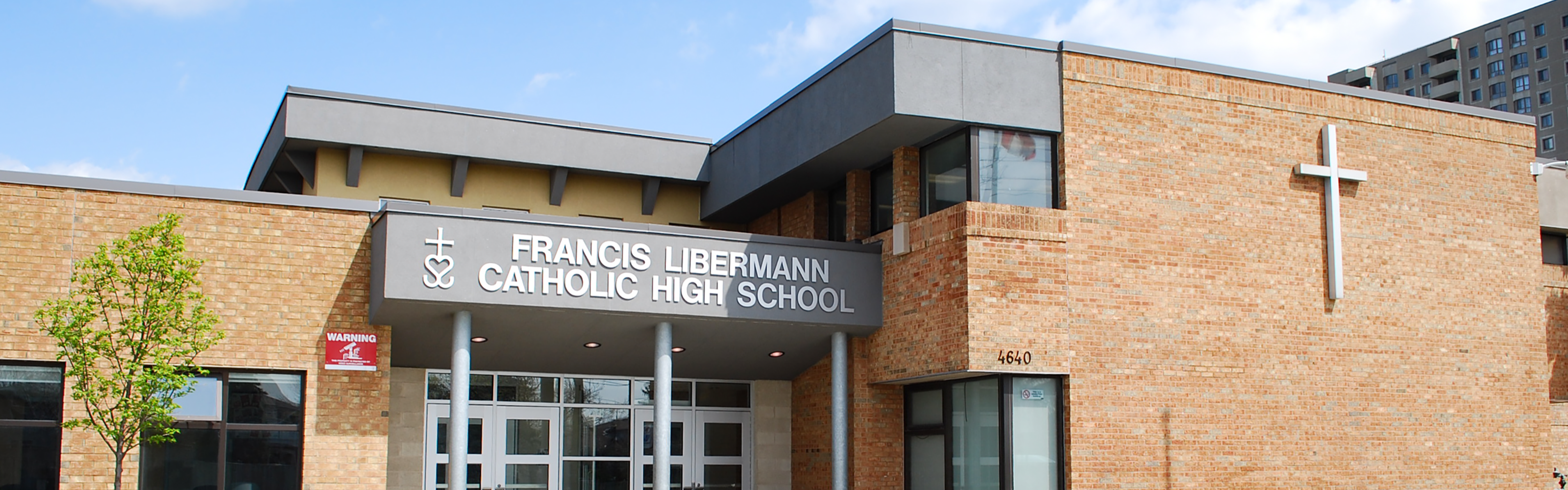 Photo of the Francis Libermann Catholic High School building, showing a red brick building with grey accents displaying the school name above the main entrance, and a large white cross on the side of the building, looking onto the road. 