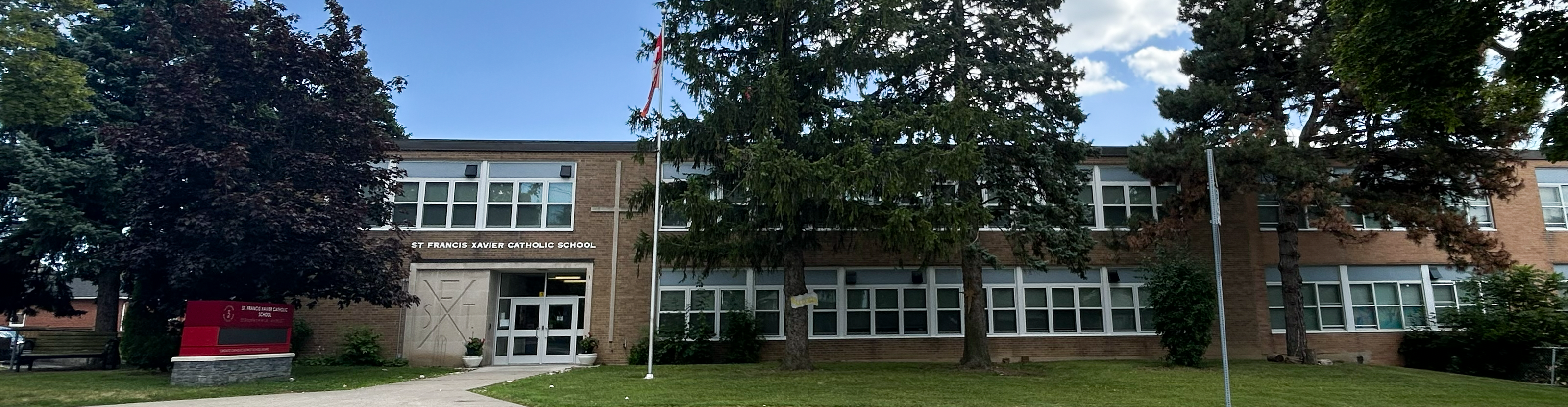 The front of the St. Francis Xavier Catholic School building, showing a beige brick building bordered by tall trees and greenery, looking out onto a residential road