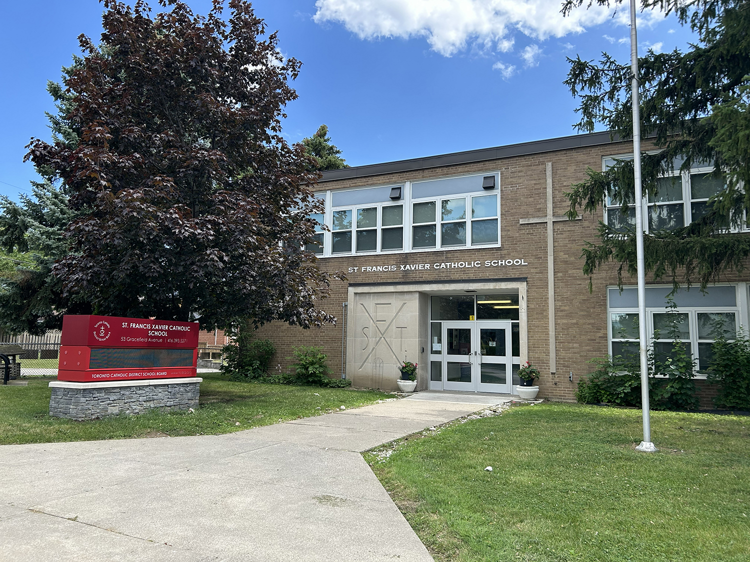 The front of the St. Francis Xavier Catholic School building, showing a beige brick building bordered by tall trees and greenery, looking out onto a residential road
