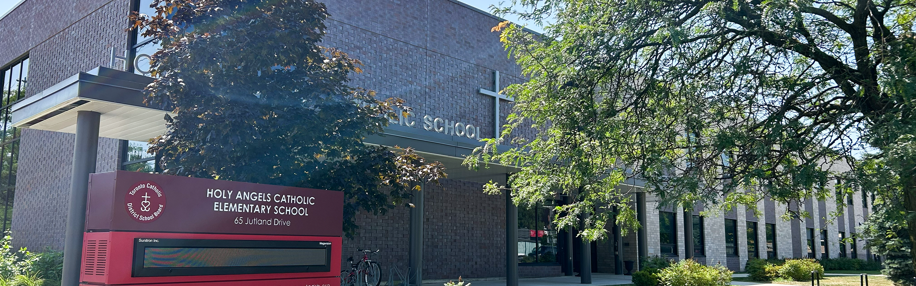 Photo of the Holy Angels Catholic School building, showing the sprawling dark and white brick building situated on the corner of a busy intersection, with verdant trees around it