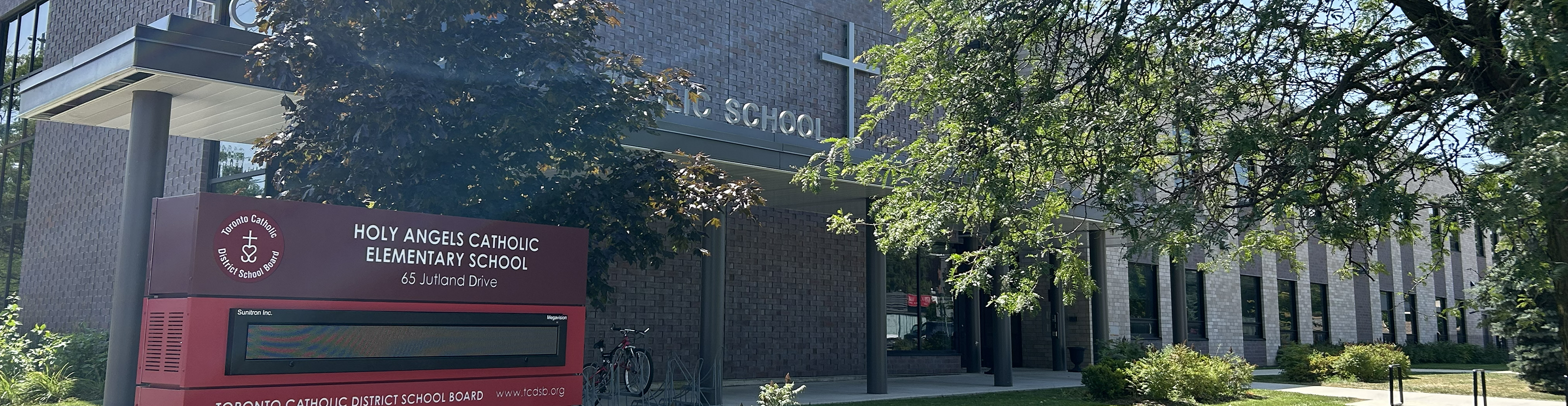 Photo of the Holy Angels Catholic School building, showing the sprawling dark and white brick building situated on the corner of a busy intersection, with verdant trees around it