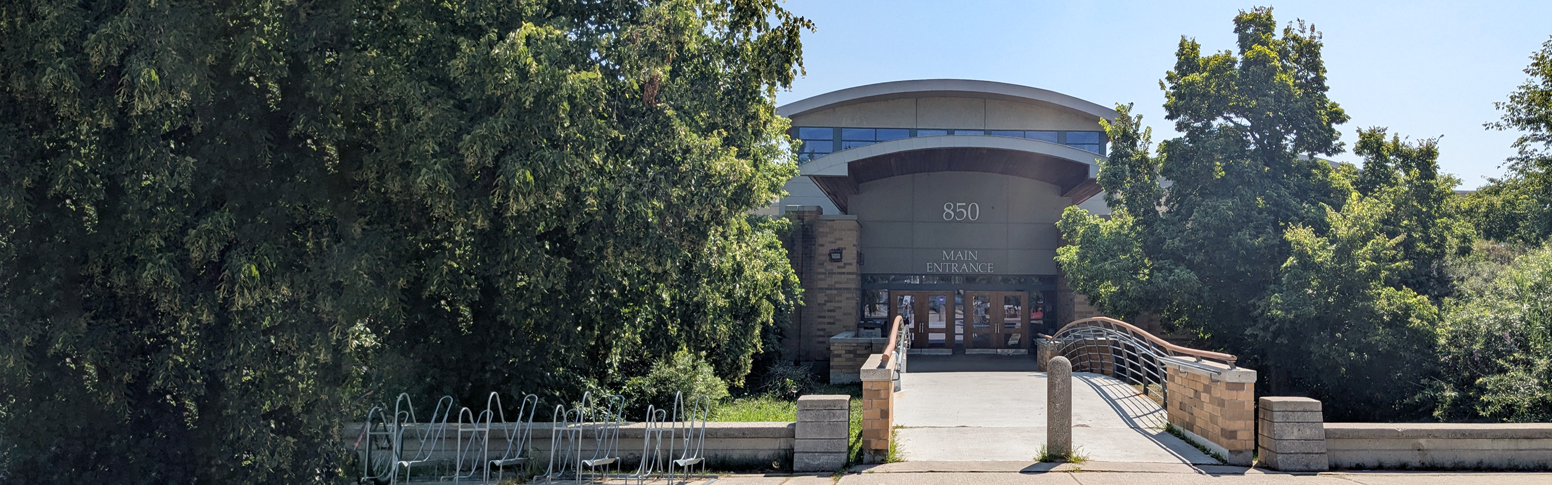 Photo of the building where the school is located, showing the main entrance of the grey and tan building, nestled between many trees, connecting the entrance to the sidewalk and bike racks with a small bridge.