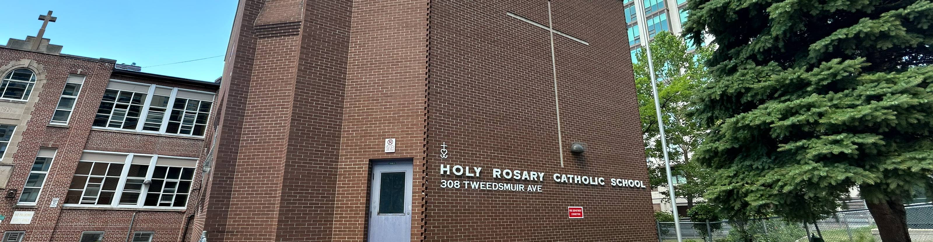 Photo of the Holy Rosary Catholic School building, showing the red and brick building nestled in a residential area, with a tall tree in front of it, and the school name and a large cross on the front of the building