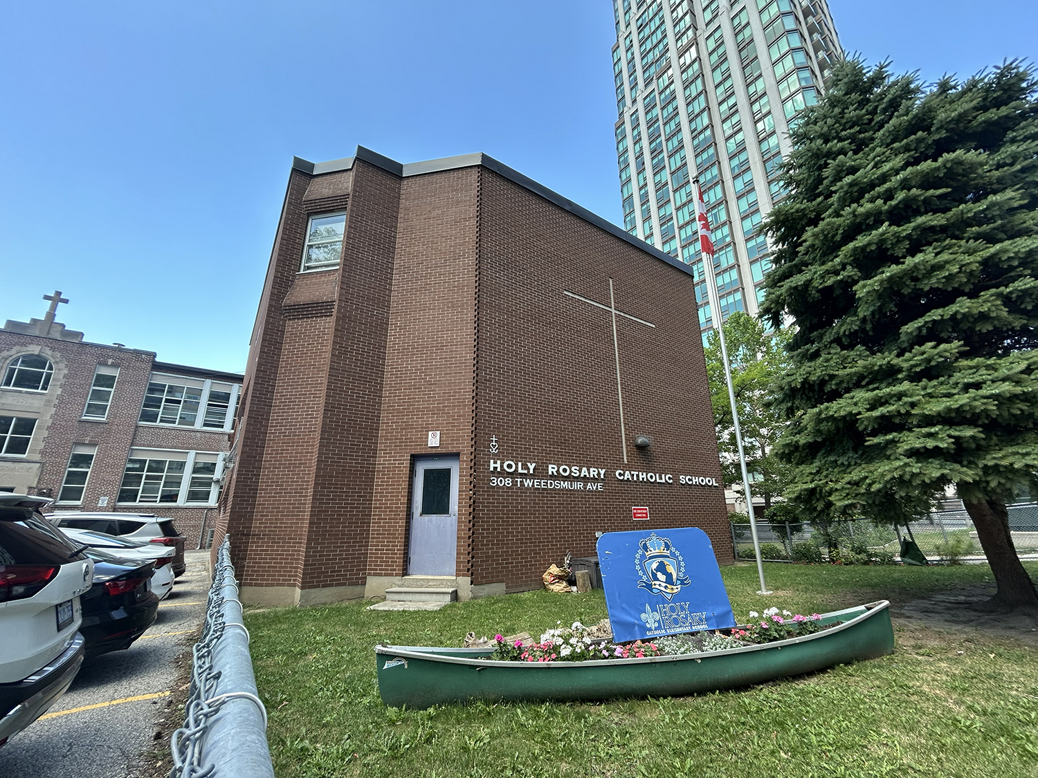 Photo of the Holy Rosary Catholic School building, showing the red and brick building nestled in a residential area, with a tall tree in front of it, and the school name and a large cross on the front of the building