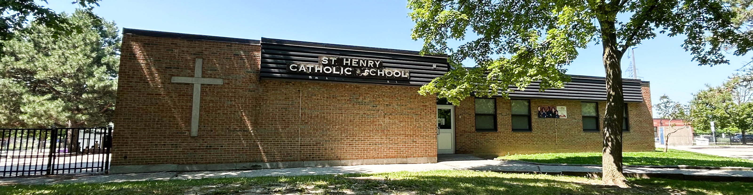 The front of the St. Henry Catholic School building, showing a light brown brick building with the school name in white on top of a black board near the top, surrounded by tall trees and green grounds