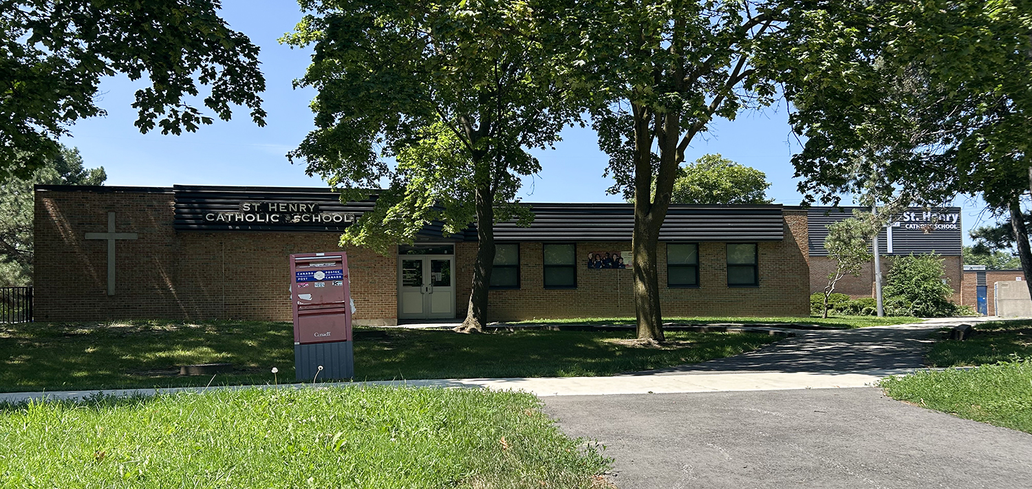 The front of the St. Henry Catholic School building, showing a light brown brick building with the school name in white on top of a black board near the top, surrounded by tall trees and green grounds
