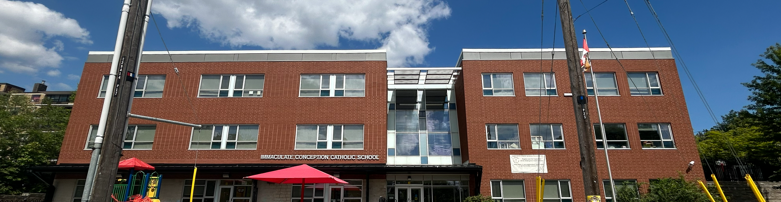 Photo of the Immaculate Conception Catholic School building, showing a red brick building with the school playground in front of it, against a blue sky