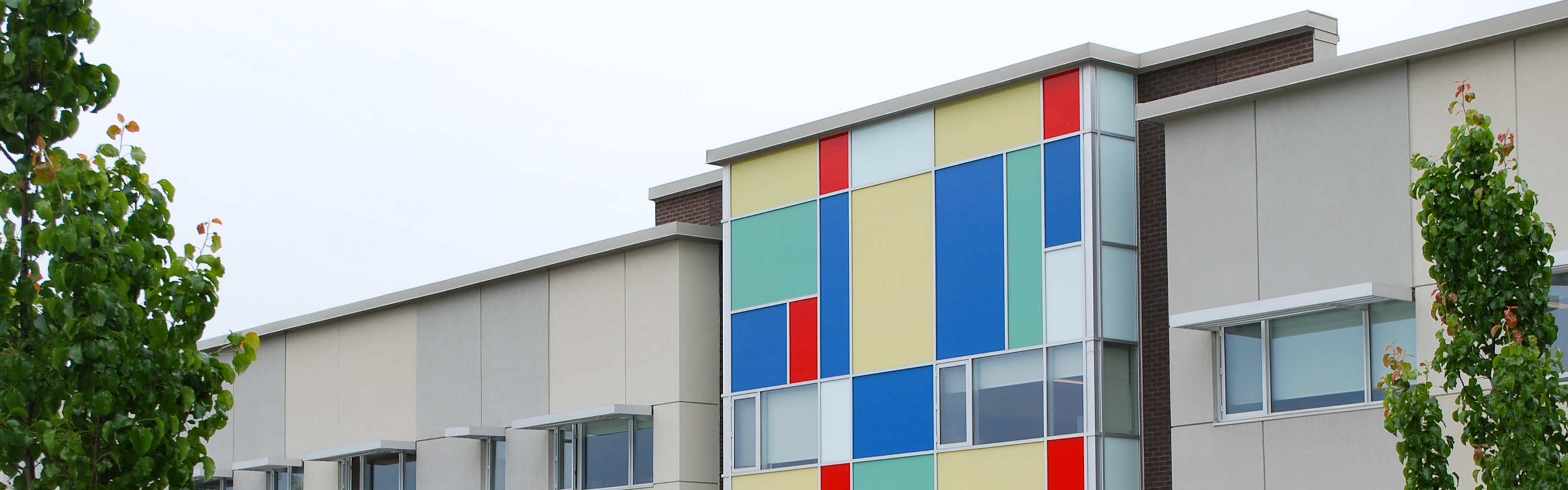 Photo of the James Cardinal McGuigan Catholic High School building, showing a modern-style brown brick and white building, with a colourful portion of the building featuring red, blue, yellow, green, and white rectangles of different sizes, framed by green trees.
